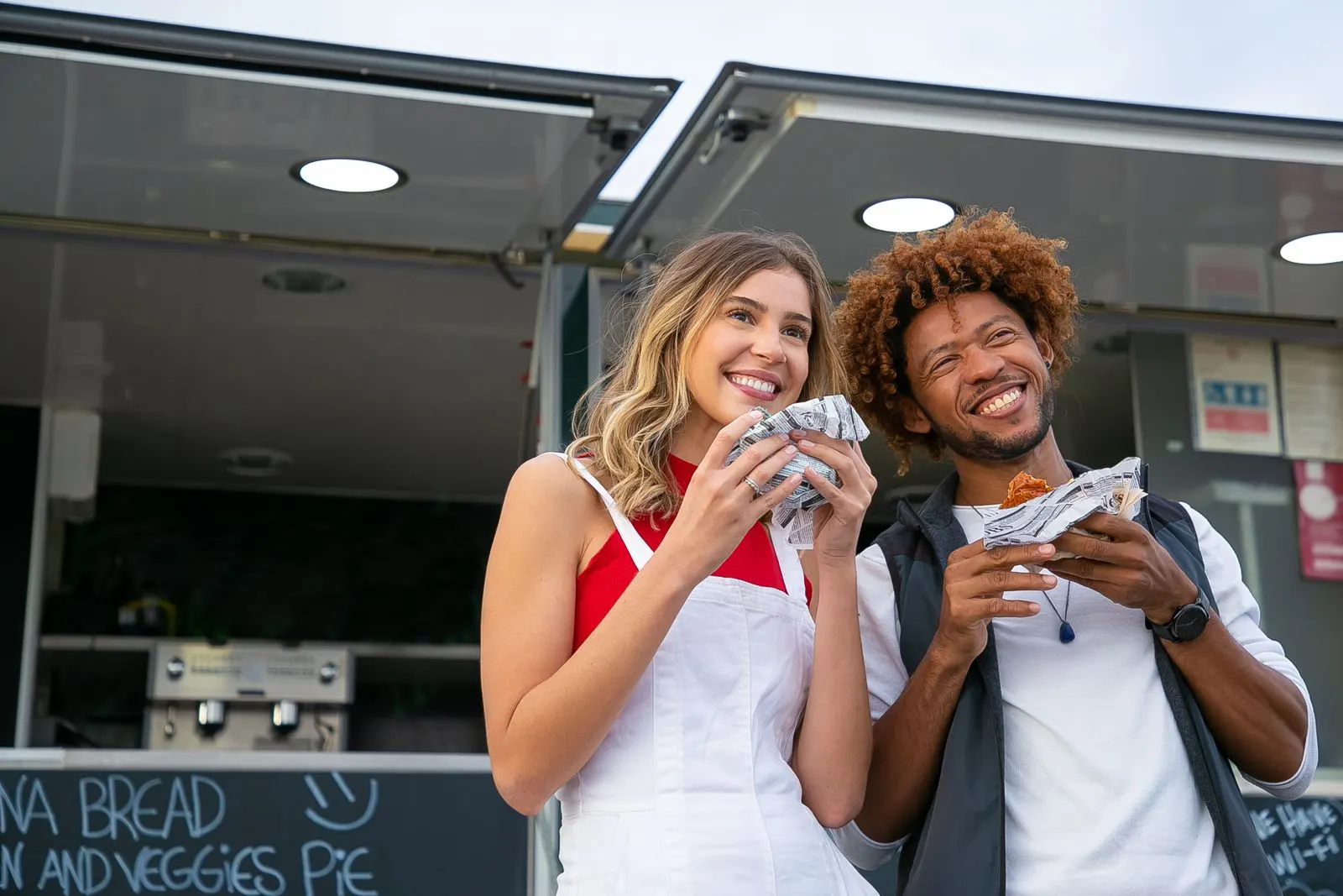 Dua teman tersenyum sambil makan burger di depan food truck